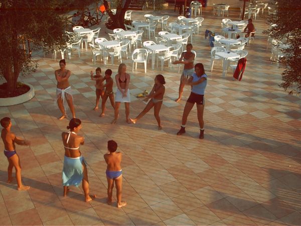 Persone partecipano a una lezione di ginnastica in una piazza con sedie bianche.