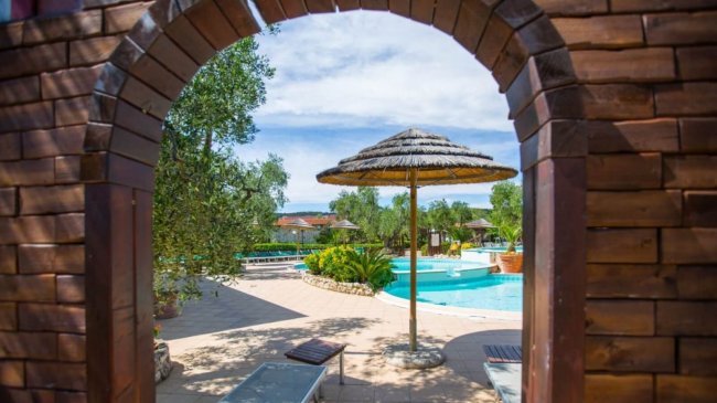 Piscina vista attraverso un arco di legno, ombrellone al centro.