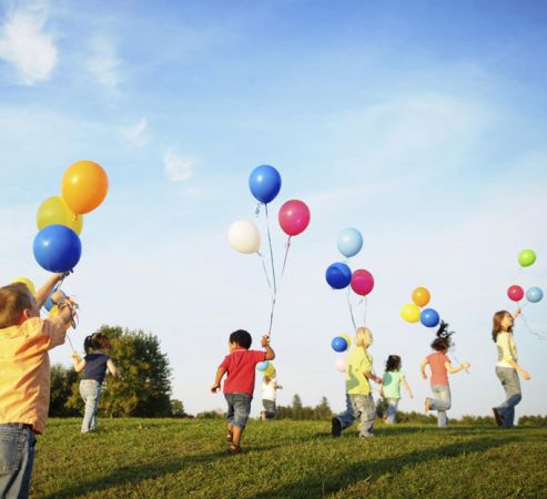 Bambini che corrono su un prato portando palloncini colorati.